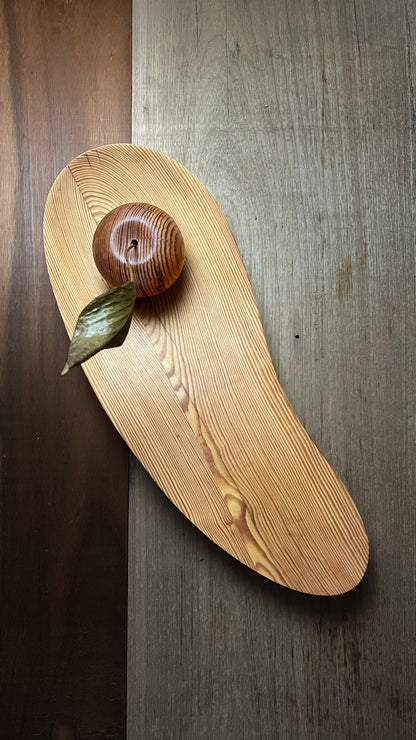 Peca autoral em Pinho de Riga - Blog Diarios do Atelier | O Designer Artesao: Travessa/bandeja decorativa de formato orgânico, esculpida em madeira, sobre a qual repousa um objeto escultórico ovoidal de madeira com detalhes de veios e uma folha seca anexada. O conjunto está disposto sobre um fundo composto por pranchas de madeira com tonalidades distintas.